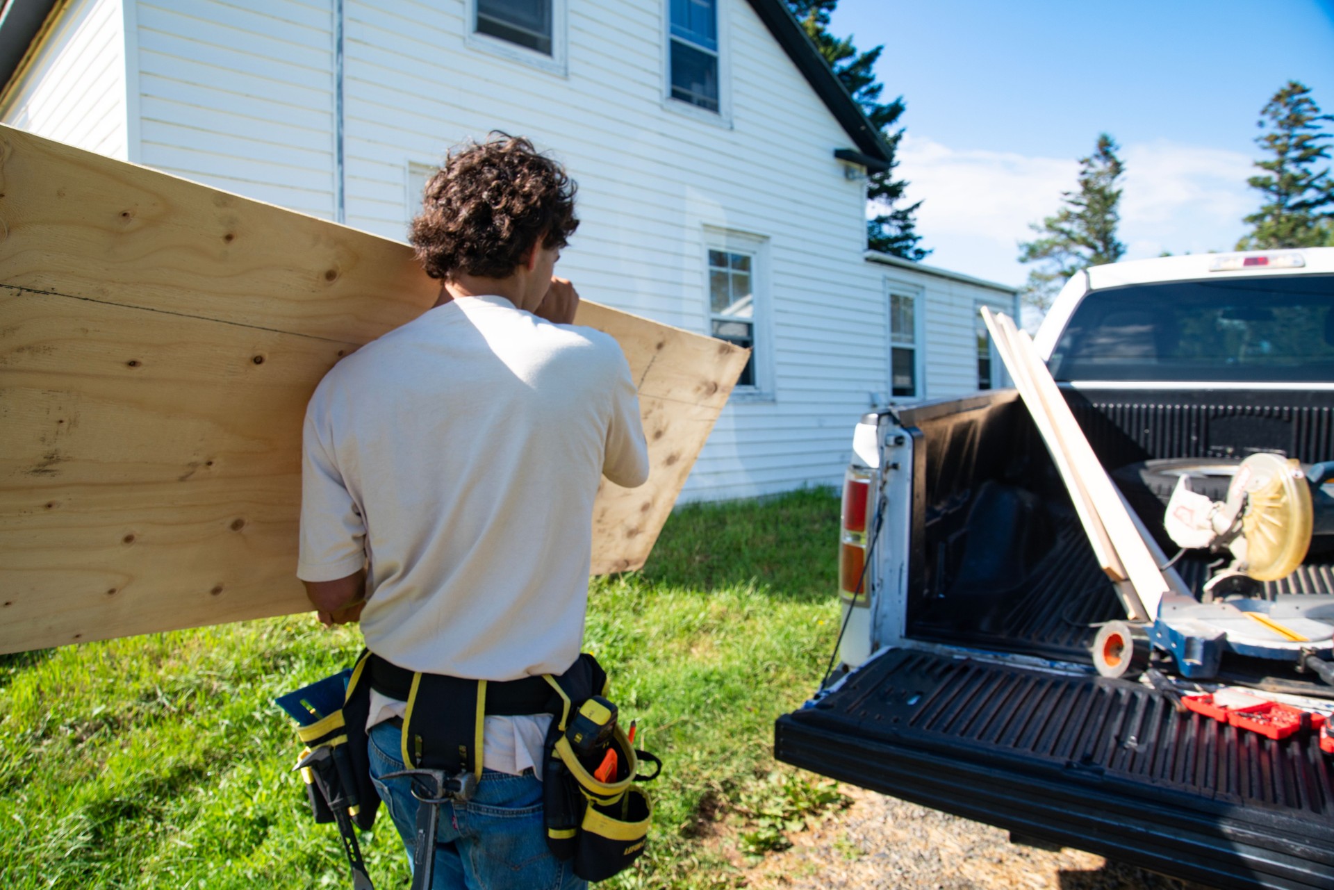 A young man carrying a sheet of plywood towards an old house he is working on.